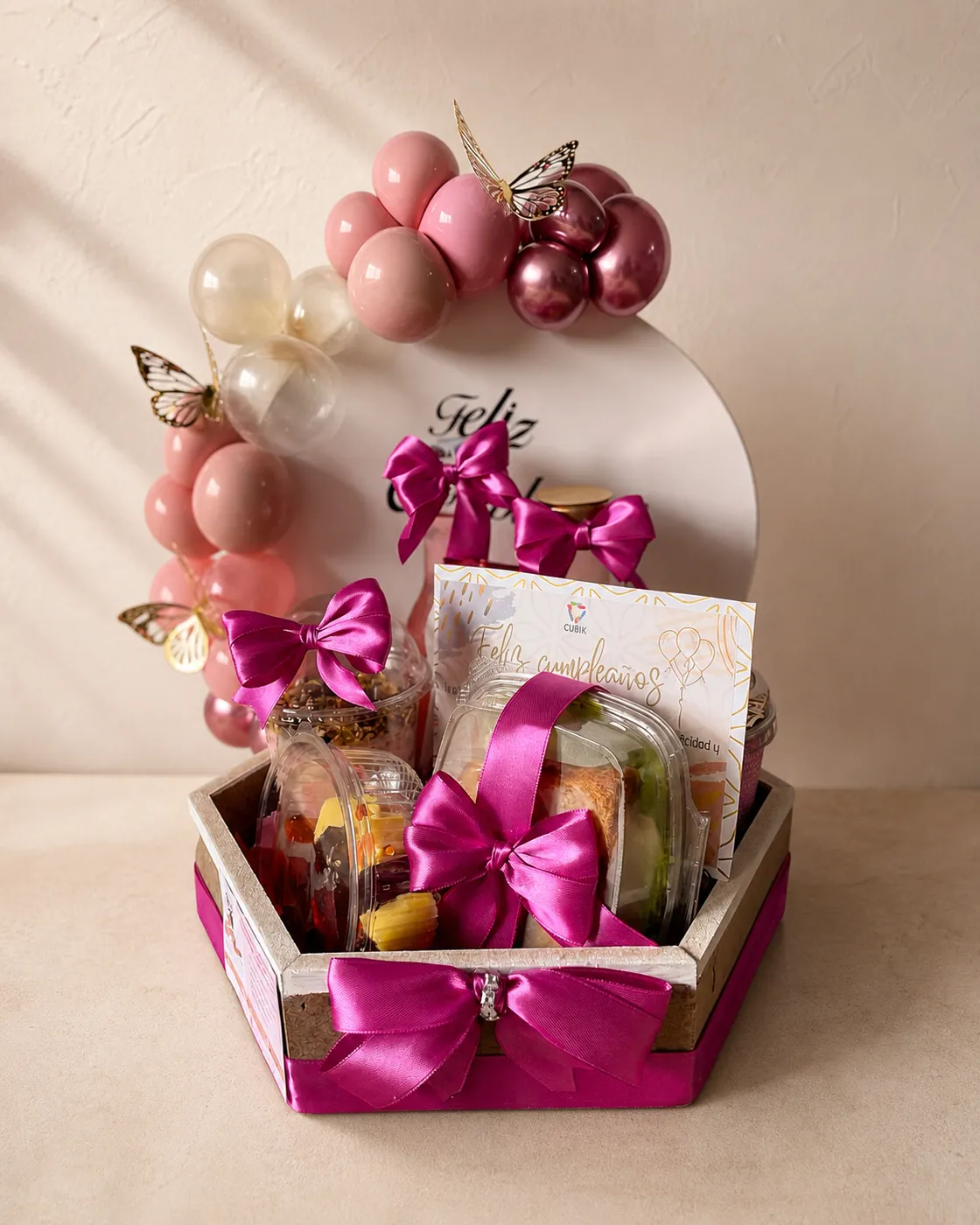 Ancheta de cumpleaños hexagonal con arco de globos rosa y granate, mariposas doradas y bandejas con fruta y aperitivos