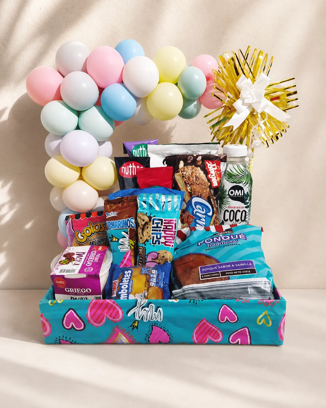 Ancheta de cumpleaños en caja turquesa con corazones, arco de globos pastel y una selección de dulces, bebidas y bizcocho